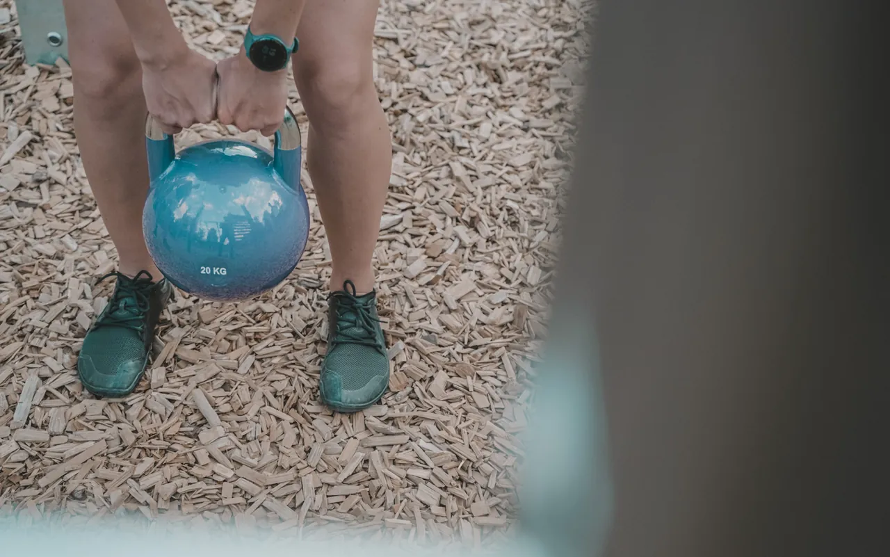 Zwei Hände greifen eine blaue Kettlebell zwischen den Füßen