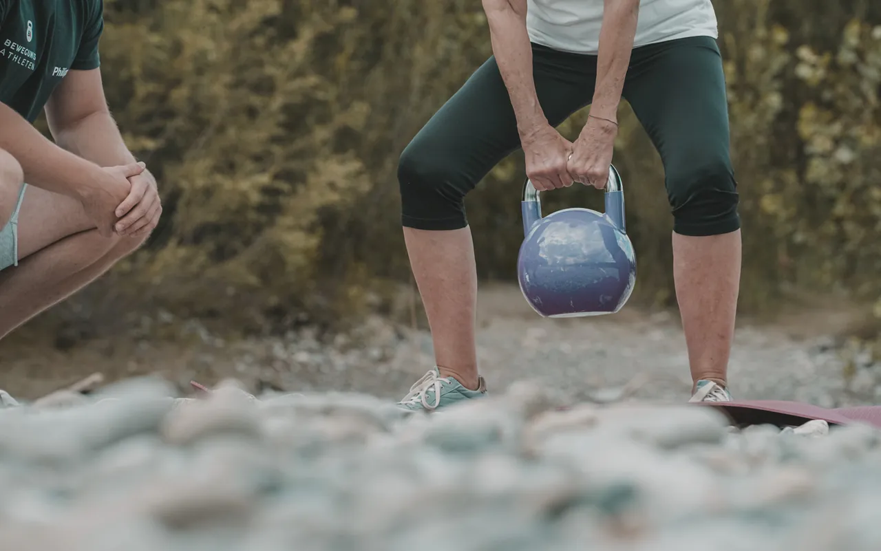 Kundin hebt eine Kettlebell vom Boden, Philipp Schmitt achtet neben ihr auf die Technik