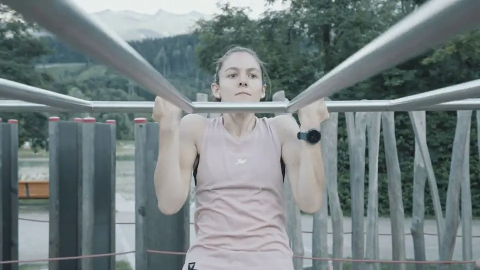 Standbild aus dem Imagefilm: Eine junge Frau hält sich im Klimmzug an einer Stange im Freien, im Hintergrund Berge