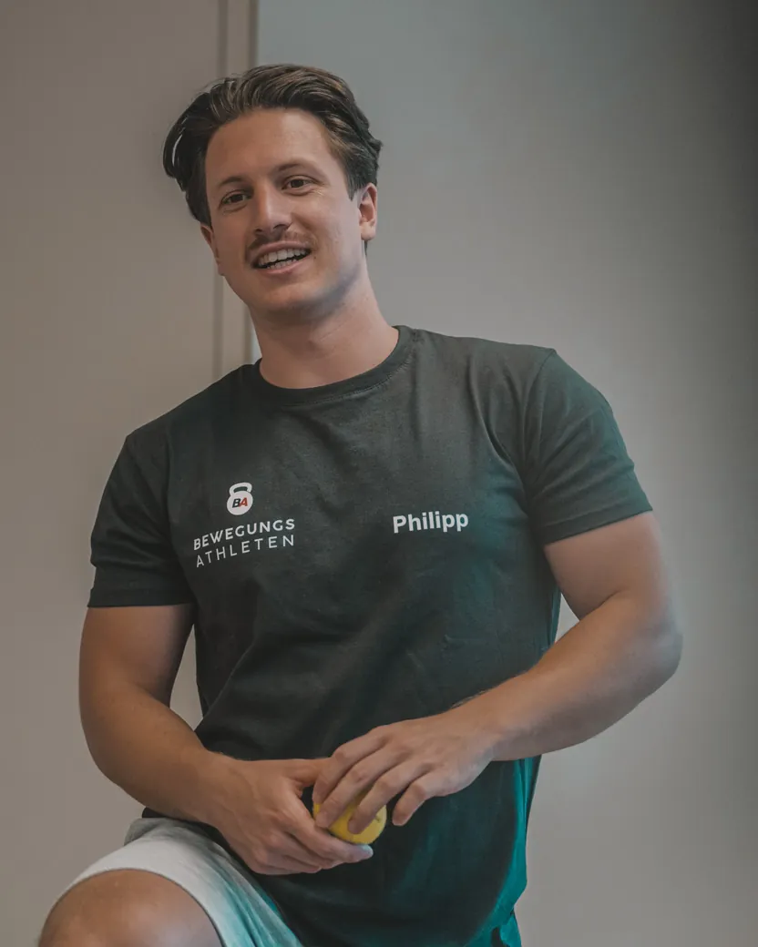 Philipp Schmitt sitzt lachend im Bewegungsathleten-Shirt und hält einen Massageball